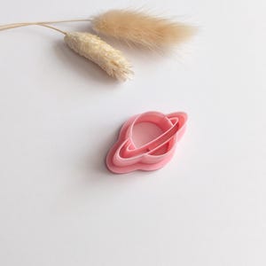 May include: A pink plastic cookie cutter in the shape of a planet with rings. The cutter is on a white surface, with dried beige decorative grass in the background. The cutter is designed for baking and crafting.
