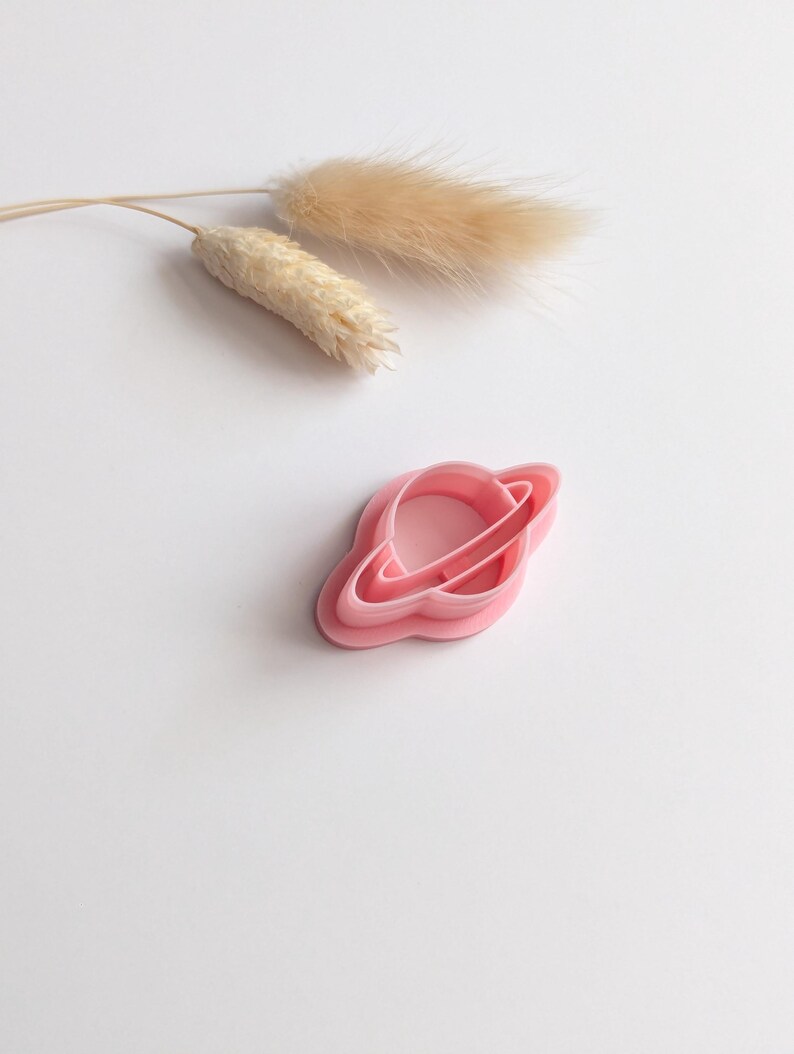 May include: A pink plastic cookie cutter in the shape of a planet with rings. The cutter is on a white surface, with dried beige decorative grass in the background. The cutter is designed for baking and crafting.