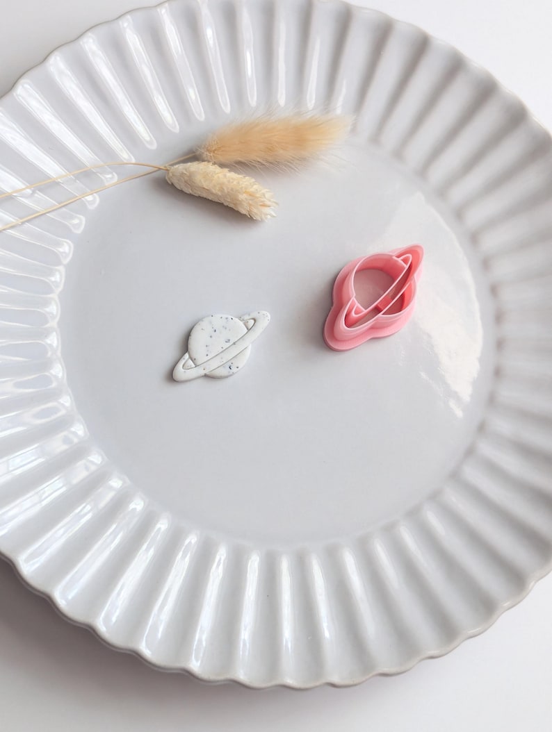 May include: A white ceramic plate with a fluted edge holds two planet-shaped items. One is white with gray speckles, the other is pink. A sprig of dried beige flowers rests on the plate.
