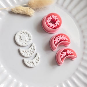 Può includere: Una collezione di strumenti per l'artigianato rosa e bianchi su un piatto bianco. Gli articoli includono un tagliapasta rotondo con un disegno a foglie, due tagliapasta a forma di mezzaluna e un tagliapasta a forma di sole. Sono presenti anche fiori beige secchi.