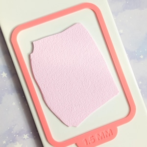 May include: A light pink, textured paper shape within a coral-coloured frame, set on a white surface. The frame has a tab with the text "1.5 MM". The background is a light cream colour.