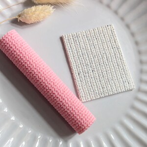 May include: A pink, textured, cylindrical object and a white, rectangular object with a knitted pattern are displayed on a white plate. Dried, beige-coloured flowers are also present, creating a minimalist aesthetic.
