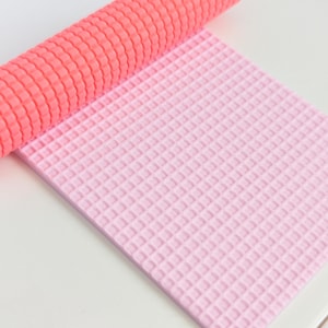 May include: A rolled coral yoga mat sits on a light pink yoga mat. Both mats have a textured, grid-like pattern. The yoga mat is designed for exercise and fitness activities.