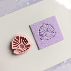 Op de afbeelding: Een roze plastic koekjesvormpje in de vorm van een schelp met een reliëf van een parel. De vorm is te zien naast een paars vierkant met een reliëf van een schelp.