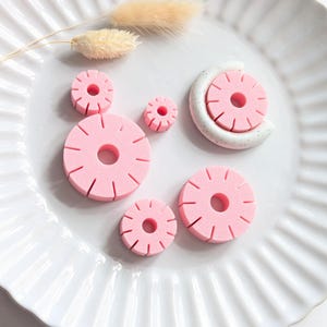 Op de afbeelding: Roze, cirkelvormige polymeerklei stukken, die lijken op miniatuur ananasschijfjes, zijn gerangschikt op een witte, geribbelde plaat. Een wit en gespikkeld halvemaanvormig stuk houdt een roze schijfje vast. Gedroogd, beige konijnenstaartgras.