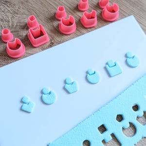 May include: A collection of pink and light blue polymer clay cutters and tools. The pink cutters are various shapes, including circles, squares, and arches. The light blue pieces are earring shapes and a textured cutting guide. The items are arranged on a white surface.