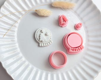 May include: A set of pink plastic cookie cutters including a snow globe shape, a circular cutter, and two small tree-shaped cutters, alongside a single gray speckled clay snow globe cutout featuring a tree and star design.