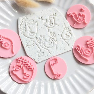 May include: A set of five circular pink stamps and one speckled white square stamp, each featuring embossed line art of abstract faces and hands. The stamps are arranged on a white fluted plate alongside two small, light-coloured dried botanical stems.