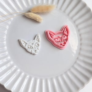 May include: Two cat-shaped cookie cutters and a decorative sprig of dried grass rest on a white, fluted plate. One cutter is pink, the other is white with speckled detailing. The cat faces are detailed with eyes, nose, and mouth.