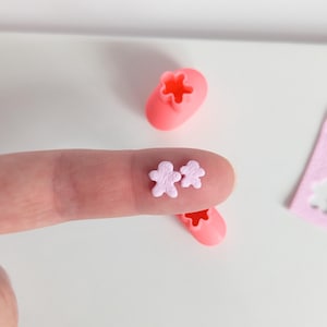 Puede incluir: Dos pequeñas chispas en forma de flor de color rosa pálido se sostienen en un dedo. También se ve un cortador de galletas en forma de flor rosa y una plantilla rosa sobre una superficie blanca. Estos elementos son para hornear o manualidades.