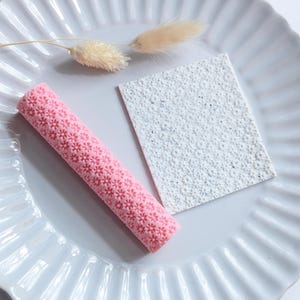 May include: A pink, flower-patterned rolling pin and a textured white square sit on a white, fluted plate. A dried, beige plant stem and flower are also on the plate. The rolling pin is cylindrical and covered in small, raised flowers.