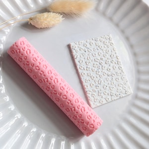 May include: A pink, cylindrical tool and a white, rectangular tool, both featuring a leopard print pattern, are displayed on a white, ridged plate. Beige dried grass adds a decorative touch. The tools are likely for crafts or baking.