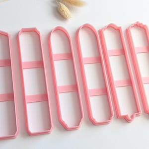 Op de afbeelding: Een set van zes lichtroze plastic koekjesvormen, elk met een lange, smalle rechthoekige vorm met verschillende boven- en onderranden, waaronder puntige, afgeronde en decoratief ingekeepte uiteinden, gerangschikt in een rij.