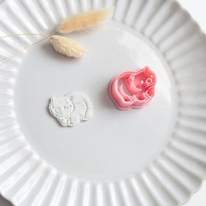 May include: A pink plastic cookie cutter shaped like a small bear and a corresponding white clay cutout with dark speckles. The bear design features a rounded body, small ears, and a smiling face. The items are arranged on a white fluted plate.