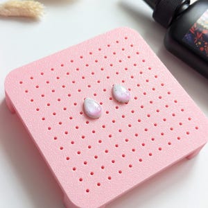 May include: Two teardrop-shaped stud earrings featuring a speckled grey base with a soft pink gradient center. The earrings are positioned on a square, light pink pegboard display stand with a grid of small holes, next to a black squeeze bottle.
