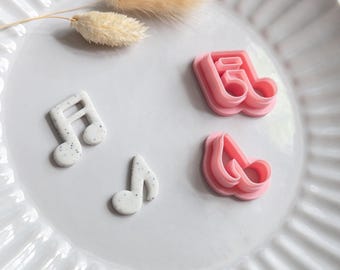 Puede incluir: Un plato blanco con borde acanalado contiene cuatro elementos con temática musical. Dos notas musicales blancas con detalles moteados y dos cortadores de galletas rosas en forma de nota musical están dispuestos en el plato, junto con una ramita de hierba seca.