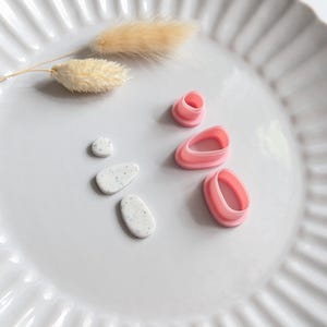 Op de afbeelding: Een witte keramische plaat met een gegolfde rand bevat knutselspullen. Twee kleine, gespikkelde, ovaalvormige kleistukken en drie roze polymeerklei-uitsteekvormen zijn op de plaat gerangschikt. Er zijn ook twee gedroogde, pluizige, beige gekleurde stelen aanwezig.