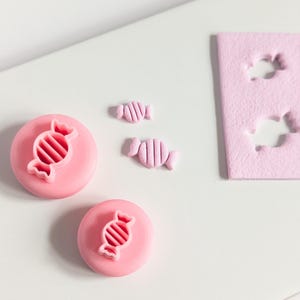 May include: Pink candy-shaped cookie cutters and candy-shaped biscuits. Two round cutters with candy designs and two small candies are next to a pink rectangular piece with candy-shaped cutouts. The items are on a white surface.