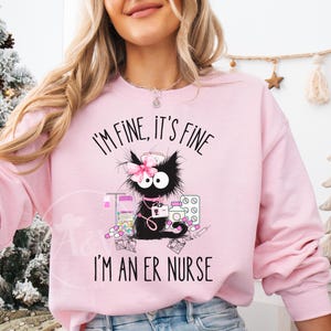 Może przedstawiać: Jasnoróżowa bluza z napisem "I'M FINE, IT'S FINE I'M AN ER NURSE" i czarnym, kreskówkowym kotem w czepku pielęgniarki. Kot otoczony jest materiałami medycznymi. W prawym dolnym rogu bluzy znajduje się napis "Light Pink".