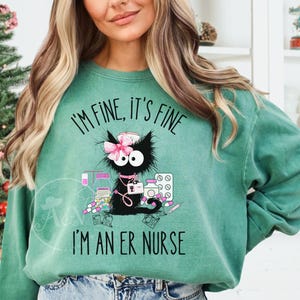 Może przedstawiać: Jasnozielona bluza z napisem "I'M FINE, IT'S FINE" i "I'M AN ER NURSE". Projekt przedstawia czarnego kota z różową kokardą i materiałami medycznymi. Bluza to wygodny, swobodny styl.