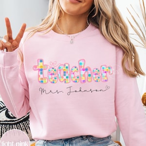 Peut inclure: Sweat-shirt rose clair avec le mot "Teacher" en lettres colorées à carreaux. Le nom "Mrs. Johnson" est écrit en dessous dans une police manuscrite. Le sweat-shirt est de style col rond.