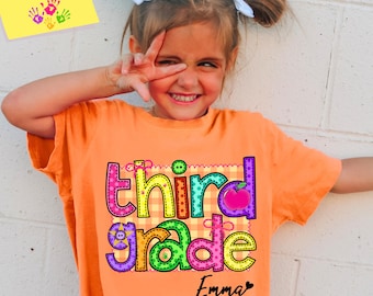 May include: An orange t-shirt with the words "third grade" in colorful letters and an apple graphic. The name "Emma" is printed below the words. A yellow sticky note in the corner says "Customization available".