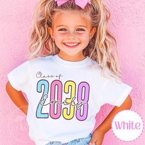 May include: A white short-sleeved t-shirt featuring the text Class of 2038 in a colourful, outlined font. The name Emily is written in a cursive script across the numbers. The numbers are coloured in light blue, pink, yellow, and purple.