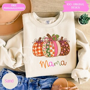 May include: A cream-coloured crewneck sweatshirt featuring a graphic of three pumpkins with various patterns, including orange plaid, floral, and polka dots, each tied with a pink bow. The word "mama" is printed below in colourful, rounded letters.