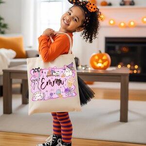 Peut inclure: Un sac fourre-tout en toile beige avec une bordure rose, portant le nom "Emma" en cursive et des illustrations sur le th&egrave;me d'Halloween. Le sac est port&eacute; par un enfant v&ecirc;tu d'un haut orange, de leggings ray&eacute;s et d'un bandeau noir et orange.