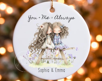 Puede incluir: Adorno de cerámica blanca con una cuerda dorada para colgar. Presenta una ilustración de dos niñas sentadas en un banco, con el texto "You + Me = Always" y los nombres "Sophia & Emma". Las niñas están rodeadas de flores.