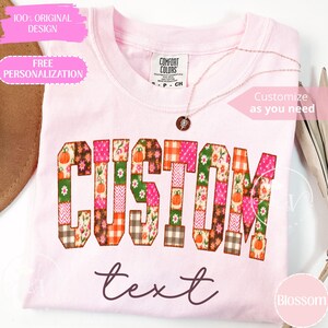 Puede incluir: Una camiseta de cuello redondo rosa claro con la palabra CUSTOM en letras grandes de bloque rellenas con un patrón de retazos de calabazas naranjas, flores y cuadros, con la palabra text escrita en letra cursiva marrón oscuro debajo.