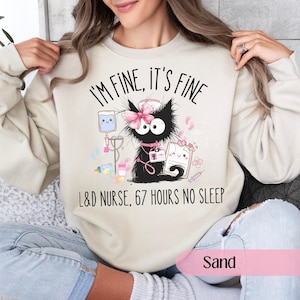 Może przedstawiać: Beżowa bluza z napisem "I'M FINE, IT'S FINE" i "L&D NURSE, 67 HOURS NO SLEEP". Grafika przedstawia czarnego kota w czepku pielęgniarki i różową kokardą, otoczonego sprzętem medycznym.