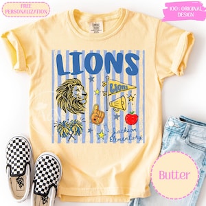 May include: A pale yellow short-sleeve t-shirt featuring a blue striped background and the word LIONS in large blue letters. The graphic includes a lion head, a pennant, a megaphone, a foam finger, pom-poms, an apple, and Jackson Elementary text.