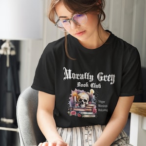 May include: A black t-shirt with a graphic design of a skull on a stack of books with candles. The text "Morally Grey Book Club" is above the skull and the text "Trigger Warnings Welcomed" is below the skull.