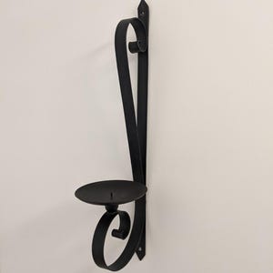 May include: Black metal wall-mounted candle holder with a scrolled design. The holder has a circular candle cup.