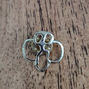 May include: A silver-coloured pin featuring a stylised figure at its centre, framed by a trefoil design. The pin has a gold-coloured centre with small, triangular accents. The pin is set against a wooden background.