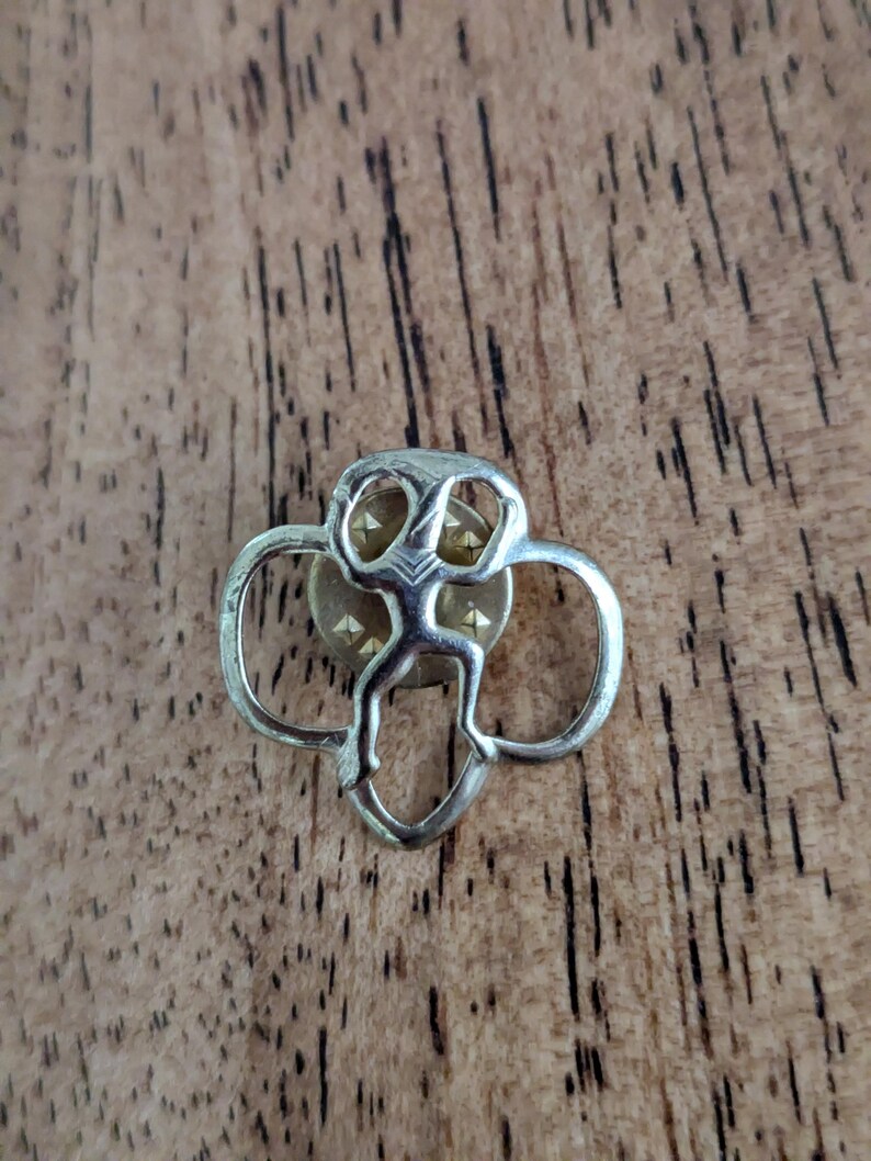 May include: A silver-coloured pin featuring a stylised figure at its centre, framed by a trefoil design. The pin has a gold-coloured centre with small, triangular accents. The pin is set against a wooden background.