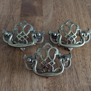 May include: Three matching brass-toned metal drawer pulls featuring an intricate openwork design with a central lattice pattern and a curved, swinging handle attached to two side mounting points on each decorative backplate.