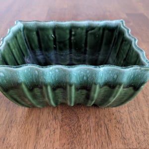 May include: A square, green ceramic planter with a scalloped rim. The planter has a ribbed texture on the sides and a glossy finish. The color is a deep, forest green with lighter green accents near the rim.