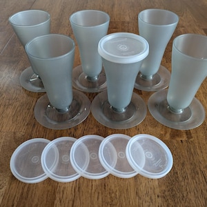 May include: A set of seven frosted glass dessert cups with clear bases and lids. The cups have a flared shape and are arranged on a wooden surface. The lids are white and round.