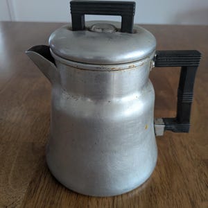 May include: Vintage aluminum coffee percolator with a black handle and lid handle. The coffee pot has a spout and a rounded body. The percolator is sitting on a wooden surface.