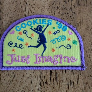 May include: A yellow patch with a purple border features the text "COOKIES '96" in turquoise, and "Just Imagine" in pink. A dark blue silhouette of a person is jumping with arms raised. Colorful swirls and stars decorate the patch.