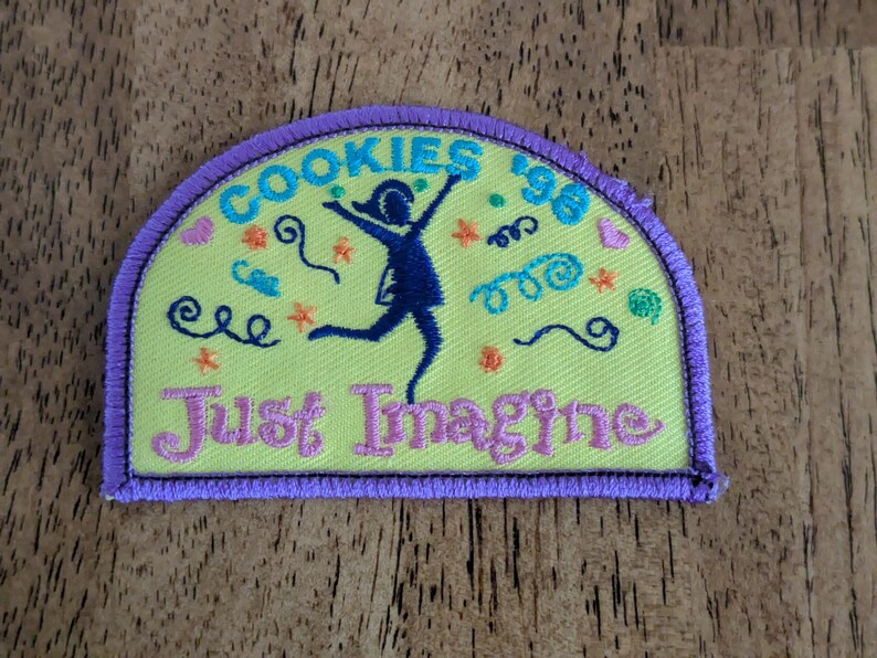 May include: A yellow patch with a purple border features the text "COOKIES '96" in turquoise, and "Just Imagine" in pink. A dark blue silhouette of a person is jumping with arms raised. Colorful swirls and stars decorate the patch.