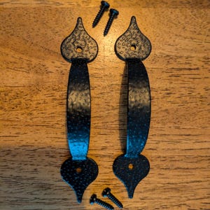 May include: Two black, textured metal door pulls with decorative teardrop-shaped ends and matching screws. The pulls have a hammered appearance and are designed for easy installation. The hardware is set against a wooden surface.