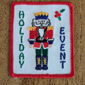 May include: Embroidered patch with a red border and a white background. The patch features a nutcracker soldier in a red coat, blue trousers, and black boots. The words "HOLIDAY EVENT" are embroidered in green and blue.