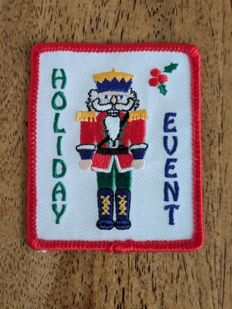 May include: Embroidered patch with a red border and a white background. The patch features a nutcracker soldier in a red coat, blue trousers, and black boots. The words "HOLIDAY EVENT" are embroidered in green and blue.