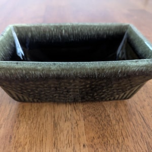 May include: A square, ceramic bowl with a textured exterior and a dark, glossy interior. The bowl has a dark olive green color with a slightly lighter rim. The bowl is sitting on a wooden surface.