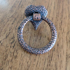 May include: A copper-toned metal drawer pull featuring a heart-shaped backplate and a circular ring handle. Both the heart and the ring have a textured, embossed surface pattern. The hardware is designed for furniture attachment.