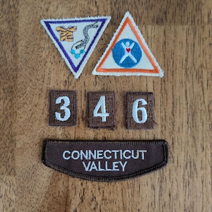 May include: A collection of embroidered patches and badges, including two triangular patches with colorful designs and three rectangular patches with the numbers 3, 4, and 6. A brown patch with the text "CONNECTICUT VALLEY" is also present.