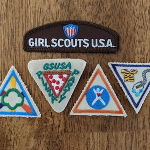 May include: Five Girl Scout badges, including a brown patch with the text "GIRL SCOUTS USA", a white badge with a pizza slice and the text "GSSUSA PIZZA PARTY", a white badge with a person with outstretched arms and a red heart, a white badge with a tiger and a snake, and a blue badge with a green clover.